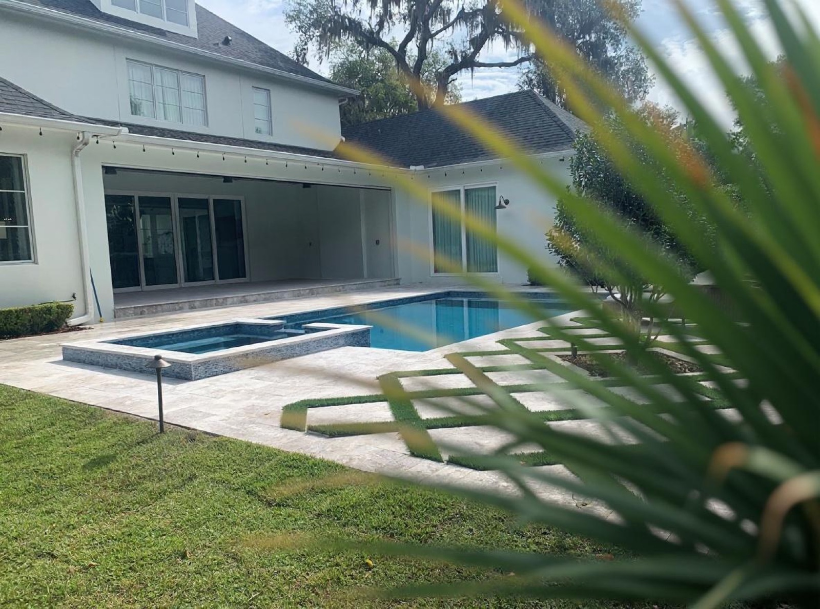 Pool and raised spa with travertine decking and geometric turf inlays, two-story white home in background